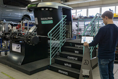 A young man stands on the right edge of the picture in front of a pedestal, pressing something on a touch display. In the background, or rather in the center of the picture, you can see a small dark staircase with a turquoise railing and a black and silver tractor cab covered in foil, in front of which is a tractor front axle. 