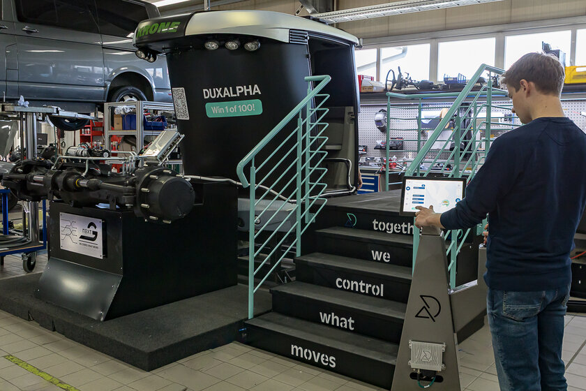A young man stands on the right edge of the picture in front of a pedestal, pressing something on a touch display. In the background, or rather in the center of the picture, you can see a small dark staircase with a turquoise railing and a black and silver tractor cab covered in foil, in front of which is a tractor front axle. 