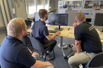 Three men are sitting in front of a PC and looking at something. The test bench can be seen in the background.