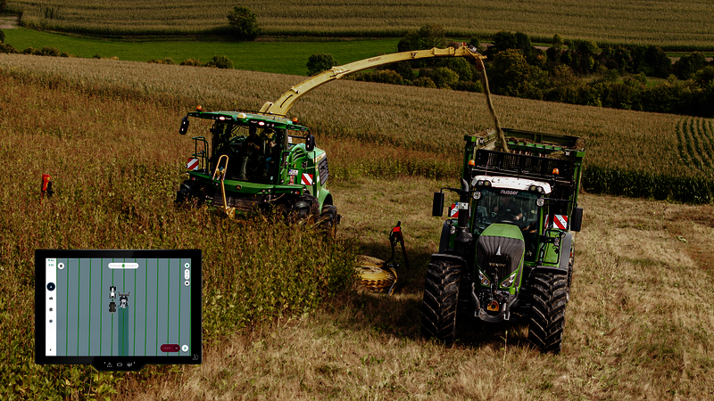 Two agricultural vehicles (a forage harvester and a tractor with a trailer) are driving across a silphie field and harvesting it. At the bottom left of the screen, you can see a touch display showing the two vehicles with track lines.