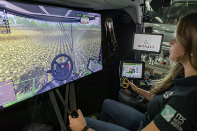 A woman is sitting in a cabin with a large screen in front of her, on which she is driving across a field. 