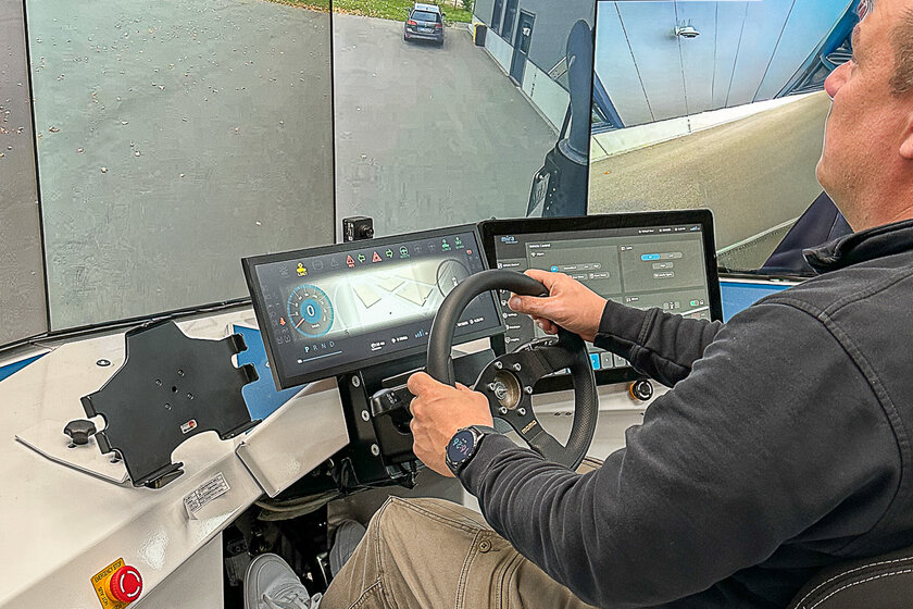 A man is sitting on the right edge of the image in front of several large screens, which can be seen on the left, showing a street. In front of the man is a steering wheel with two touch displays.