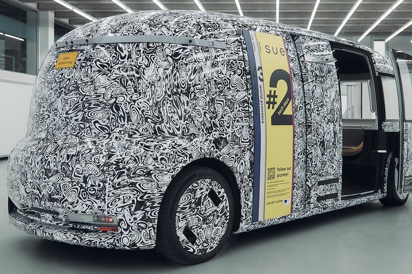 A bus that is still camouflaged (black and white pattern) stands in a sparsely lit, bright hall. The bus has no windows.