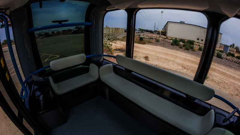 The interior of a vehicle with windows all around and white benches in front of them. This vehicle does not have a steering wheel. There is space to stand in the middle.