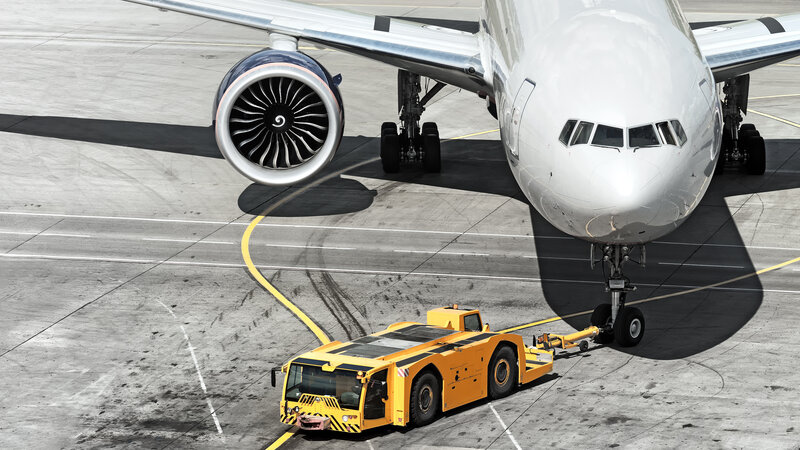 On an airport runway, a so-called pusher, i.e. a yellow aircraft tractor, pulls a large aircraft from the top right to the front left.