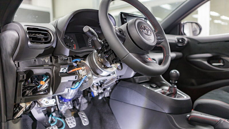 A car cockpit that no longer has a steering column on the steering wheel, but instead has a motor mounted there. In addition, a few orange, turquoise, and dark blue cables hang from the floor, connecting to another motor. 