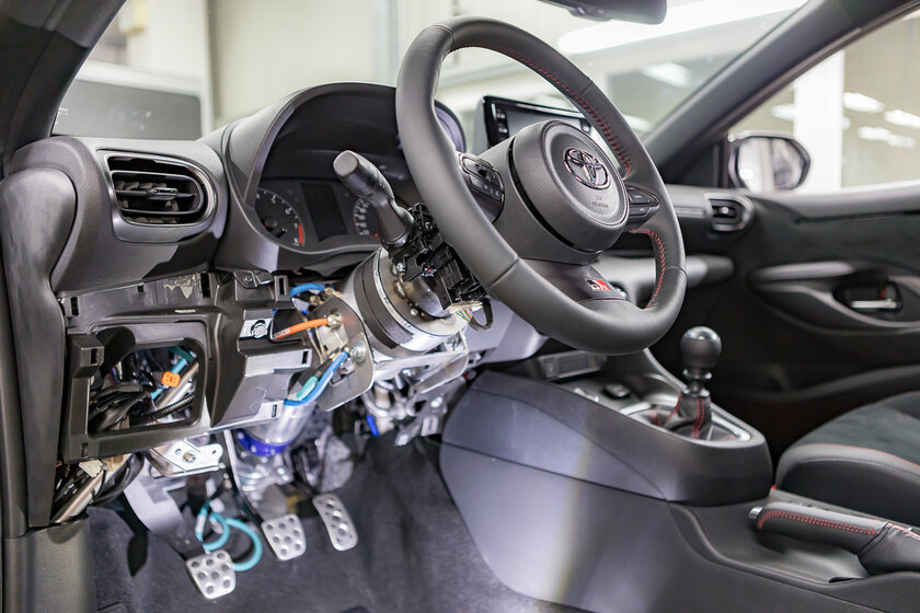 A car cockpit that no longer has a steering column on the steering wheel, but instead has a motor mounted there. In addition, a few orange, turquoise, and dark blue cables hang from the floor, connecting to another motor. 