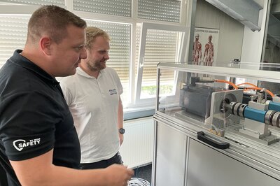 Two male figures look from the left to the right at a test stand in an office, smiling slightly.