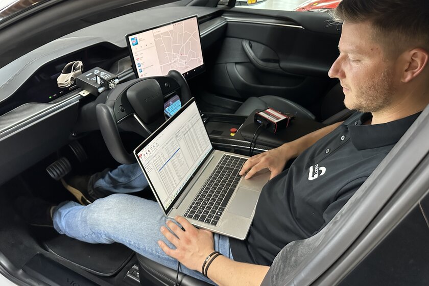 A man is sitting in a car with his laptop in front of him, running a software program. Next to him and in front of him are various gateways and cables.