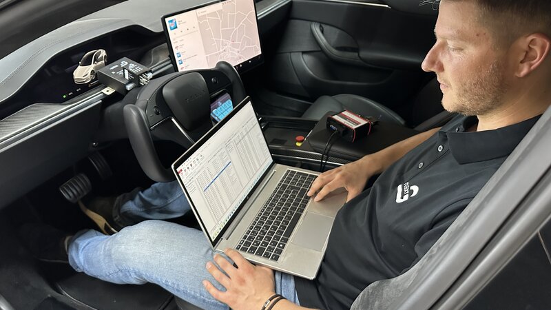 A man is sitting in a car with his laptop in front of him, running a software program. Next to him and in front of him are various gateways and cables.