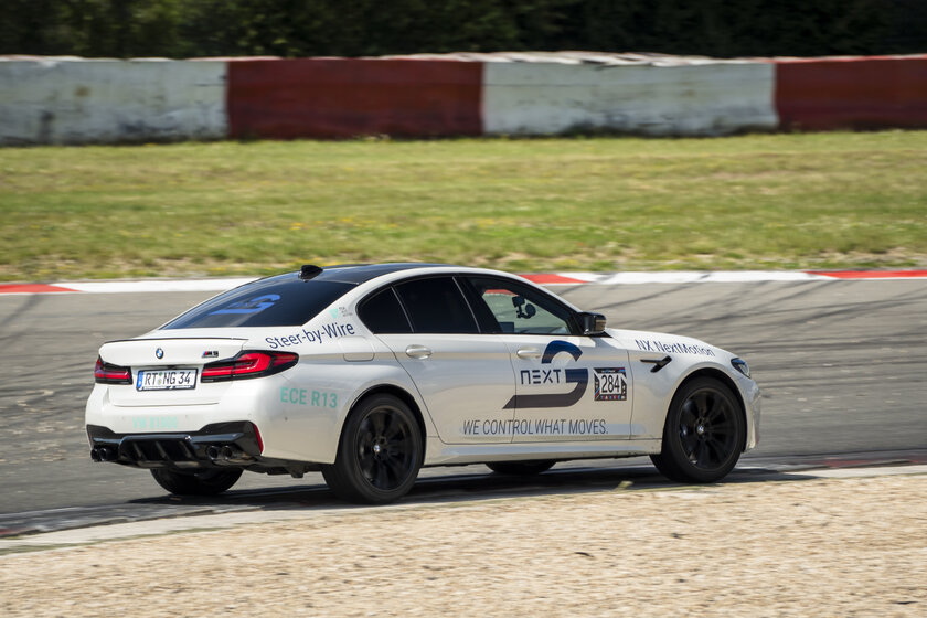 A white BMW is driving pretty fast on a racetrack. The car is adorned with various logos, including the main Arnold NextG logo.
