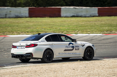 A white BMW is driving pretty fast on a racetrack. The car is adorned with various logos, including the main Arnold NextG logo.