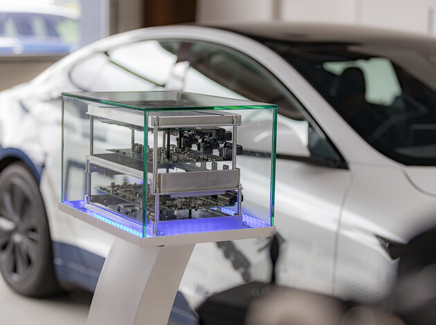 evels so that the circuit boards can be seen inside. The whole thing is presented in a glass display case. A white Tesla stands blurred in the background.