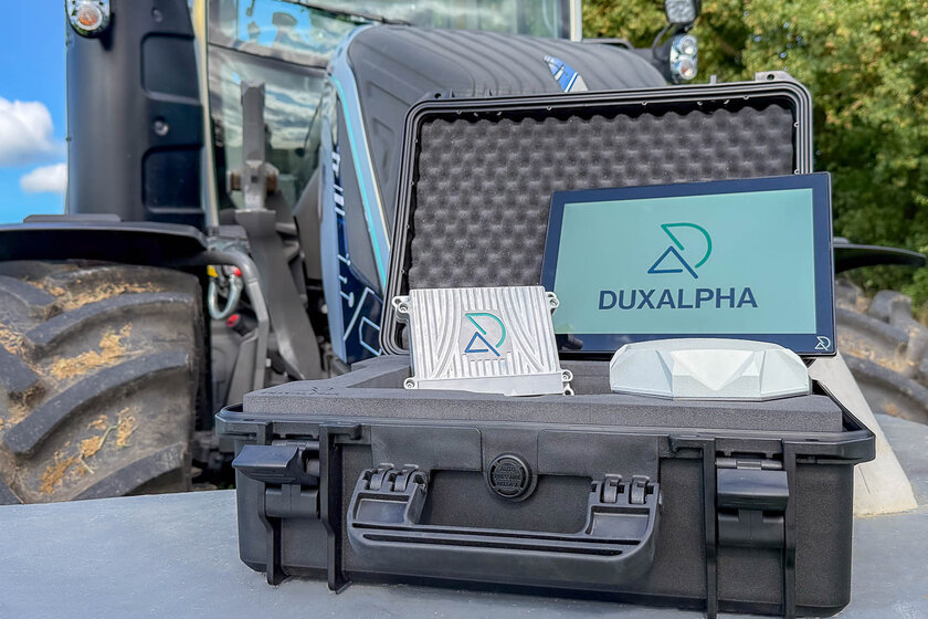 A tractor is blurred in the background. In the foreground is an open black suitcase containing a touch display with the DuxAlpha logo on it. To the left of it is a DuxAlpha-branded ECU and in front of it is a white antenna.
