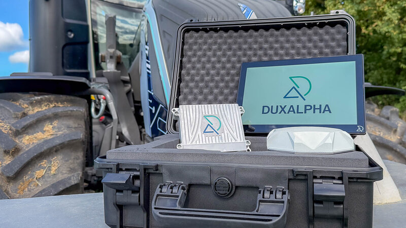 A tractor is blurred in the background. In the foreground is an open black suitcase containing a touch display with the DuxAlpha logo on it. To the left of it is a DuxAlpha-branded ECU and in front of it is a white antenna.
