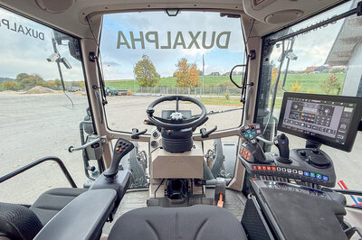 You see the interior of a tractor from the driver's position. You look out through the windshield, where “DuxAlpha” is written in large letters, upside down.