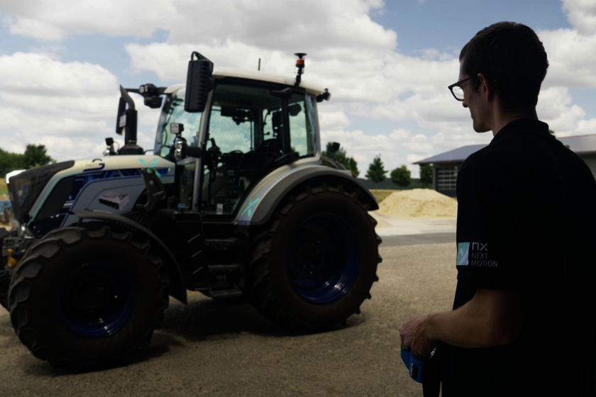 A man stands in front of a silver-blue tractor with no one sitting in it. The man visible on the right edge of the picture stands with a remote control in his hand and controls the tractor.  