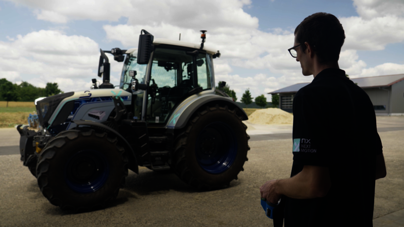 A man stands in front of a silver-blue tractor with no one sitting in it. The man visible on the right edge of the picture stands with a remote control in his hand and controls the tractor.  