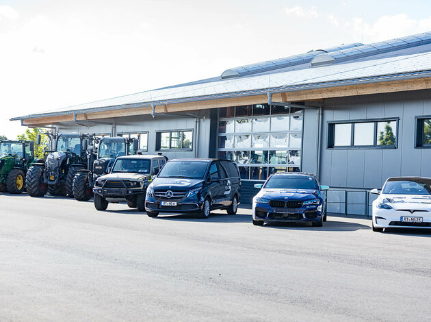 Tractors, off-road vehicles, vans, and cars – including Mercedes, BMW, and Tesla – parked in front of a modern industrial building with a garage door.