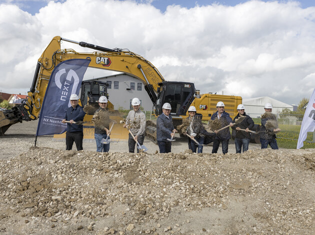 Several people, including Kevin and Luca Arnold, stand behind a pile of dirt with a shovel and throw the dirt forwards. On the left is an Arnold NextG flag, on the right a PARAVAN flag and in the background a large tracked excavator.