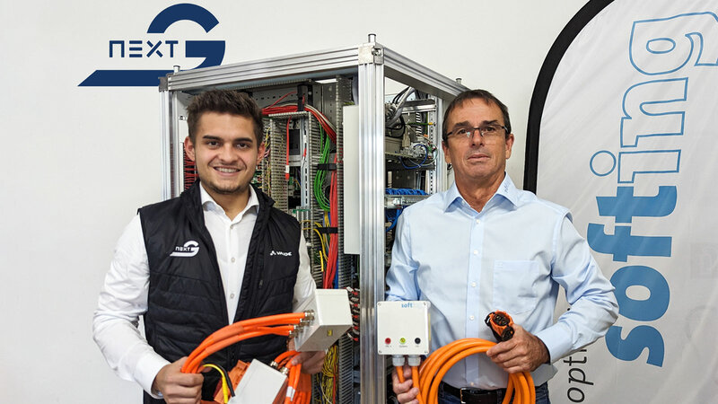 Two men are standing next to each other, each holding an orange cable. In the background is a large technical cabinet with lots of cables and a softing beach flag.