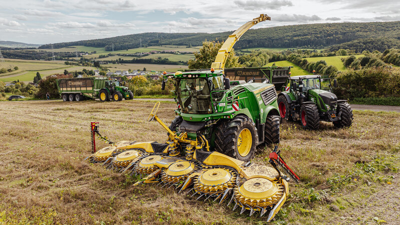 Three agricultural vehicles are standing in a field. At the front is a green and yellow chopper, behind it are two more tractors, each with a large trailer.