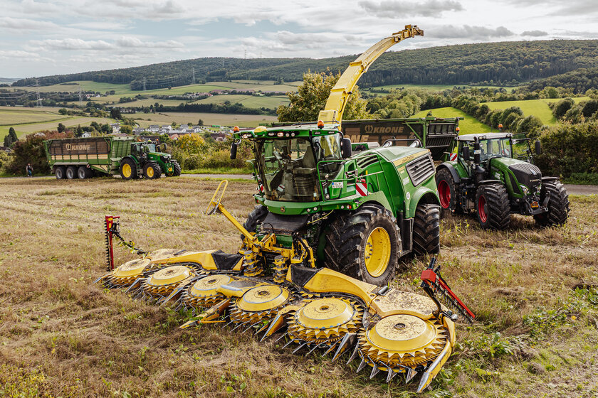Three agricultural vehicles are standing in a field. At the front is a green and yellow chopper, behind it are two more tractors, each with a large trailer.