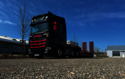 A black and red Scania S 770 with a red and grey low-loader is standing on a gravel car park next to a hall under a bright blue sky.