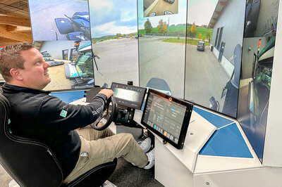 A man sits in front of a steering wheel with several large screens in front of him. The screen shows a panoramic view of a vehicle.