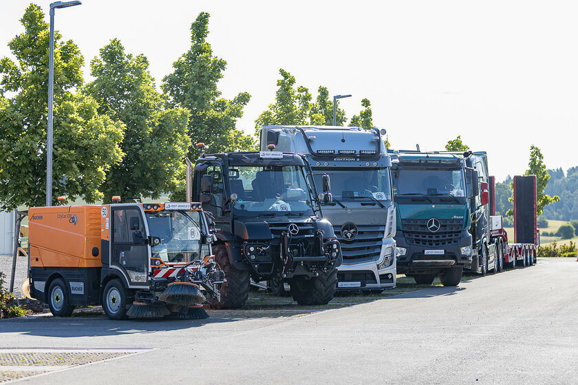A lineup of various commercial vehicles, including a street sweeper, Unimog, and multiple Mercedes-Benz trucks, parked side by side in front of trees.
