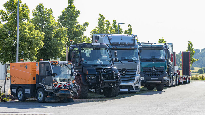 A lineup of various commercial vehicles, including a street sweeper, Unimog, and multiple Mercedes-Benz trucks, parked side by side in front of trees.