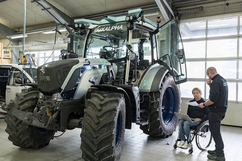 A tractor is standing in a hall. To the right of the tractor are two people who are discussing something. One of the two men is sitting in a wheelchair.