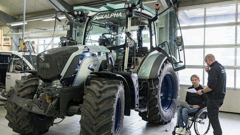 A tractor is standing in a hall. To the right of the tractor are two people who are discussing something. One of the two men is sitting in a wheelchair.