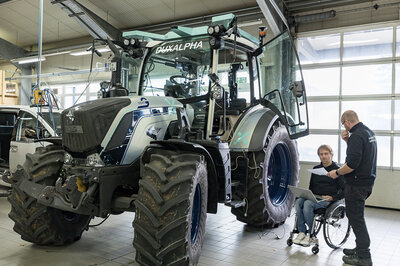 A tractor is standing in a hall. To the right of the tractor are two people who are discussing something. One of the two men is sitting in a wheelchair.