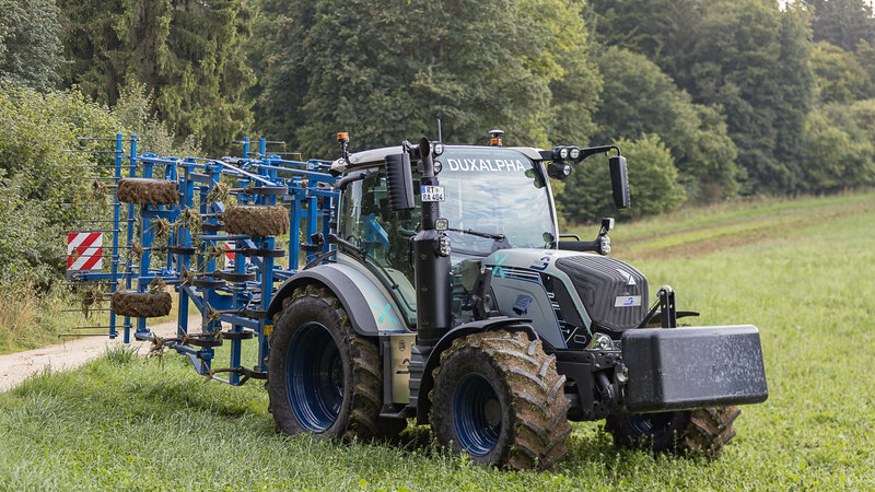 A silver-blue tractor with a blue attachment is standing on a meadow. The tires and the attachment are covered in soil.