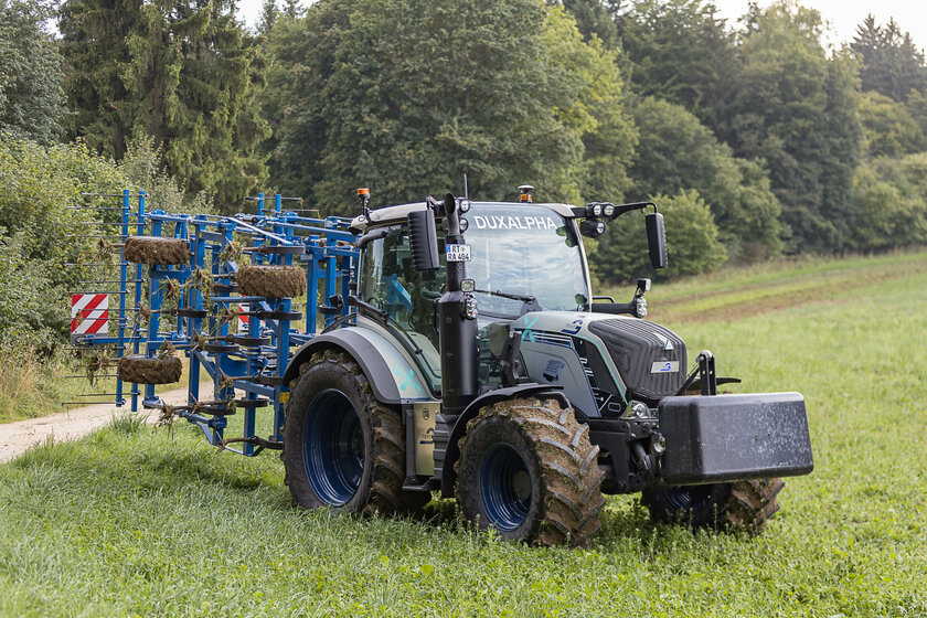 A silver-blue tractor with a blue attachment is standing on a meadow. The tires and the attachment are covered in soil.