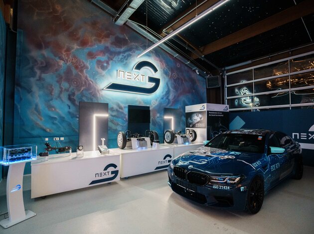 A rather dark room in which a lot of blue can be seen. On the right is a blue BMW M5, on the left is an illuminated Arnold NextG logo on the wall and underneath are tables with exhibits and a television.