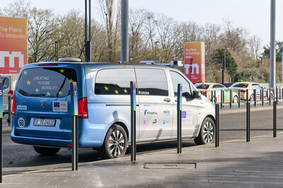 A blue and silver vehicle is parked at the side of a road next to bollards. A camera system for autonomous or teleoperated driving is mounted on the car.