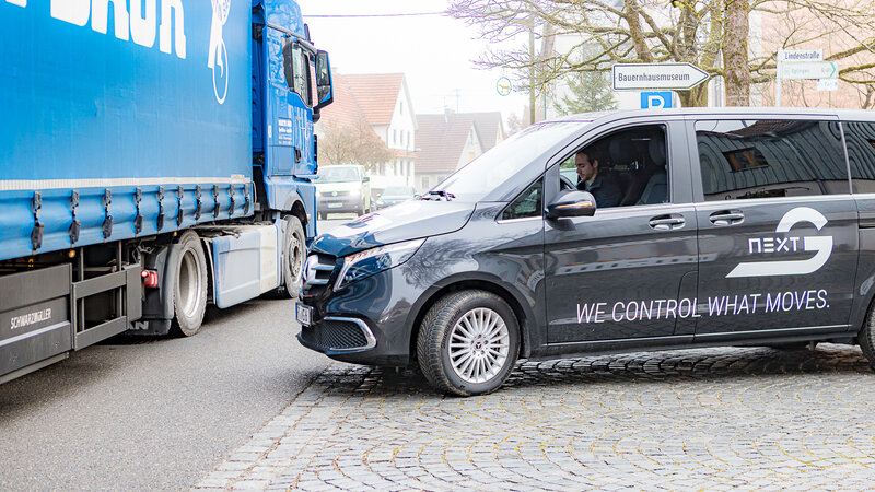 A blue truck drives past on the left side of the road. A V-Class van covered with Arnold NextG logos approaches from the right and looks to the left. There is no one in the driver's seat, only a man in the passenger seat looking at his cell phone.