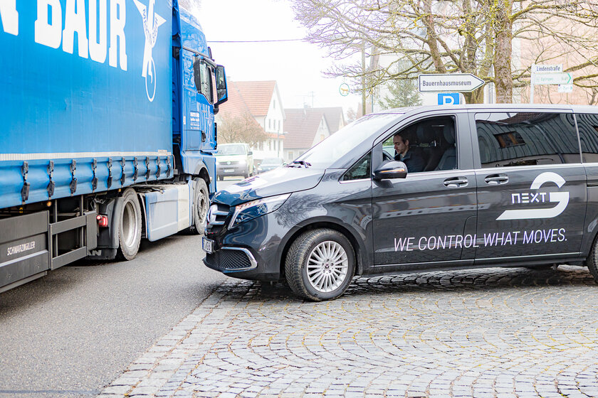 A blue truck drives past on the left side of the road. A V-Class van covered with Arnold NextG logos approaches from the right and looks to the left. There is no one in the driver's seat, only a man in the passenger seat looking at his cell phone.