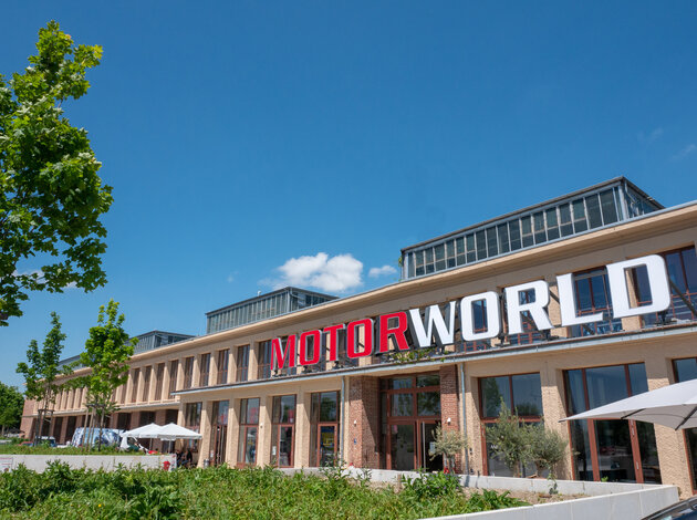 Location Munich. Entrance to Motorworld Munich, a long, rather flat, slightly red building, with a blue sky and sunshine.