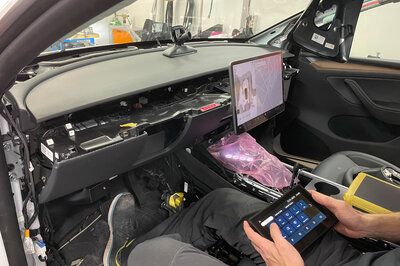 The interior of a car that is undergoing conversion. There is no longer a steering wheel, and a person is sitting in the driver's seat holding a touch display for various functions in the car.