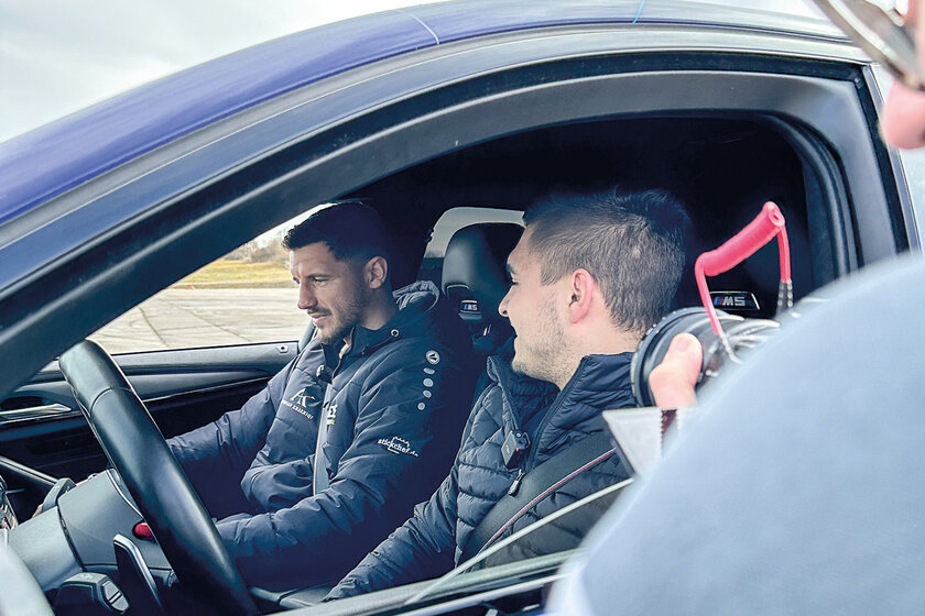Two brown-haired young men are sitting in a car and talking to each other. Another person stands in front of the lowered driver's window and films into the car.