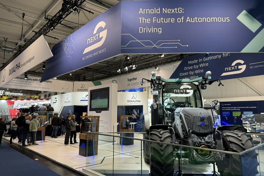On the right edge of the image is a silver tractor with a large NextG logo on the windshield. The tractor is located at our booth at Agritechnica 2025, with a white floor, blue banners with text on the ceiling, and a large TV in the background.