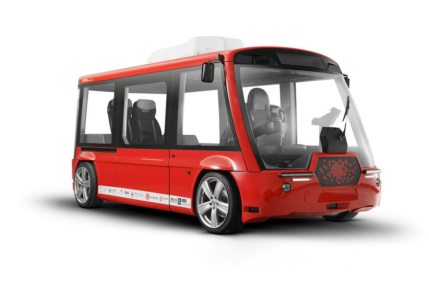 A small red bus with a new interior design. The driver does not have a steering wheel, only a joystick on the right-hand side. There are two additional seats in the rear of the bus.