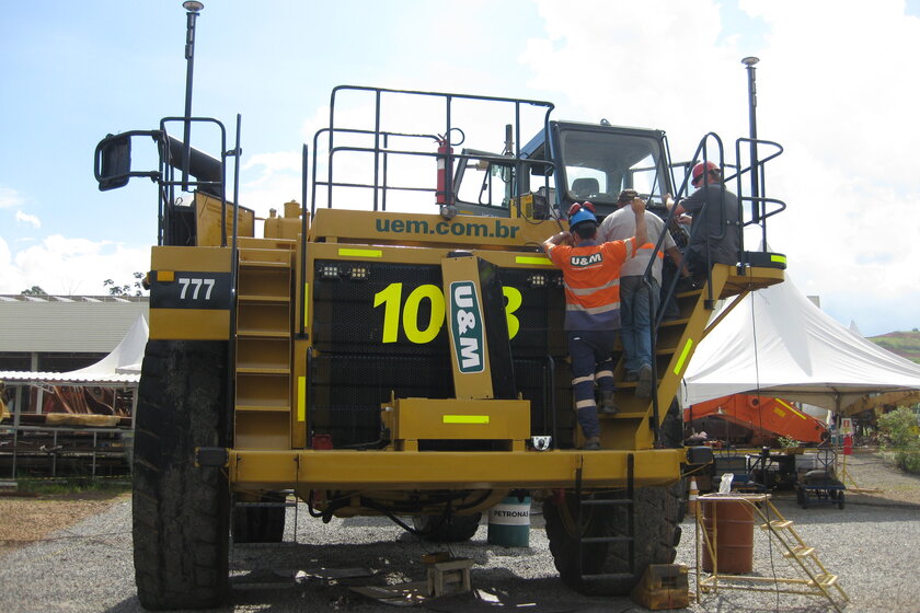 A yellow dump truck that is currently undergoing renovation. Three people are standing on the ladder leading up to the cab of the dump truck, which does not have a dump body due to the renovation work.
