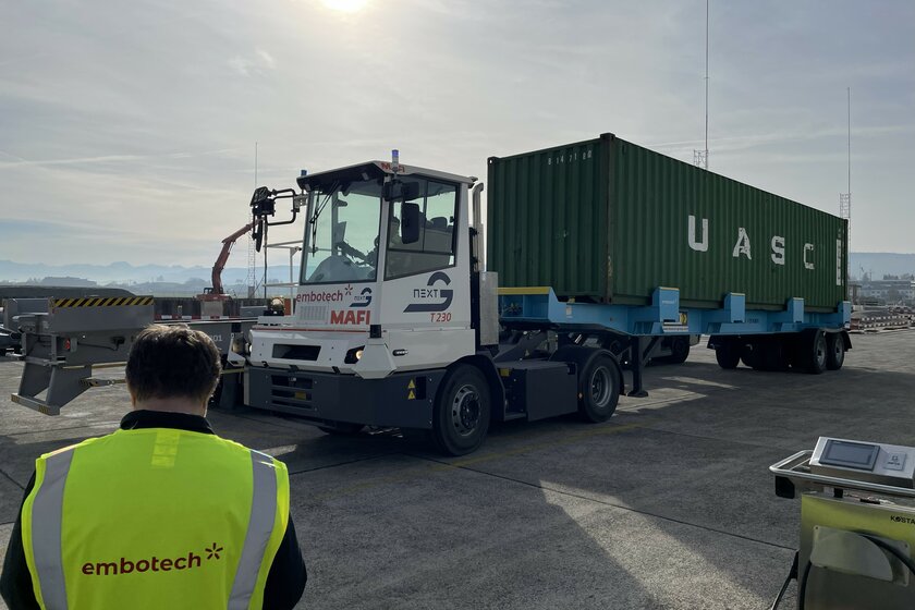 A white and gray terminal truck with a green container trailer attached to the rear, standing in the sunshine on a concrete yard.