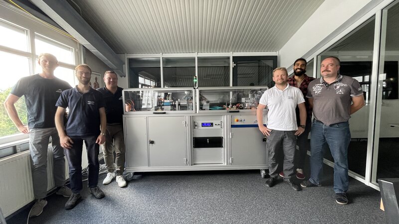 Three people each stand to the left and right of a shoulder-high 2m long box in an office.