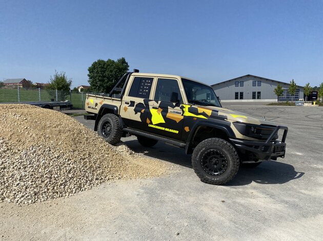 Modified Toyota Landcruiser with camouflage wrap and off-road tires positioned in front of a gravel pile on a test site.