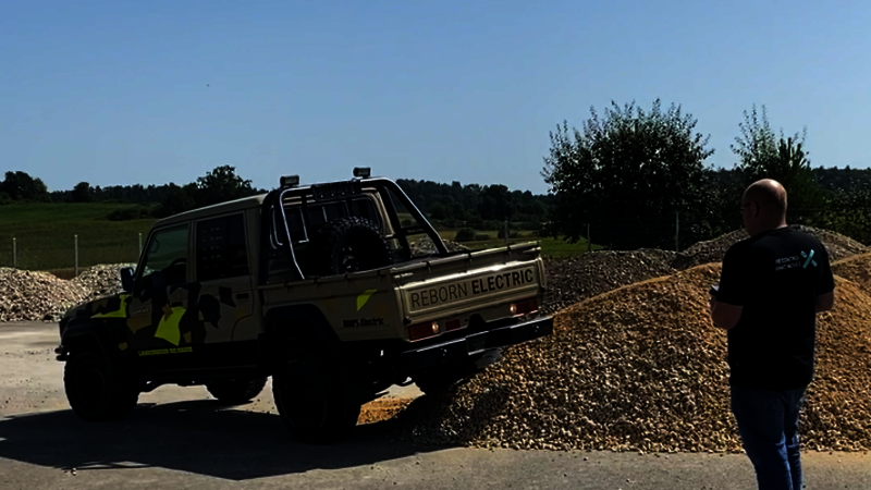 A Toyota Landcruiser with double cab for underground mining in brown with black and neon-colored highlights, drives with the right rear wheel on a pile of gravel, controlled by an Arnold NextG employee who looks at the Toyota and has a remote control in his hand.